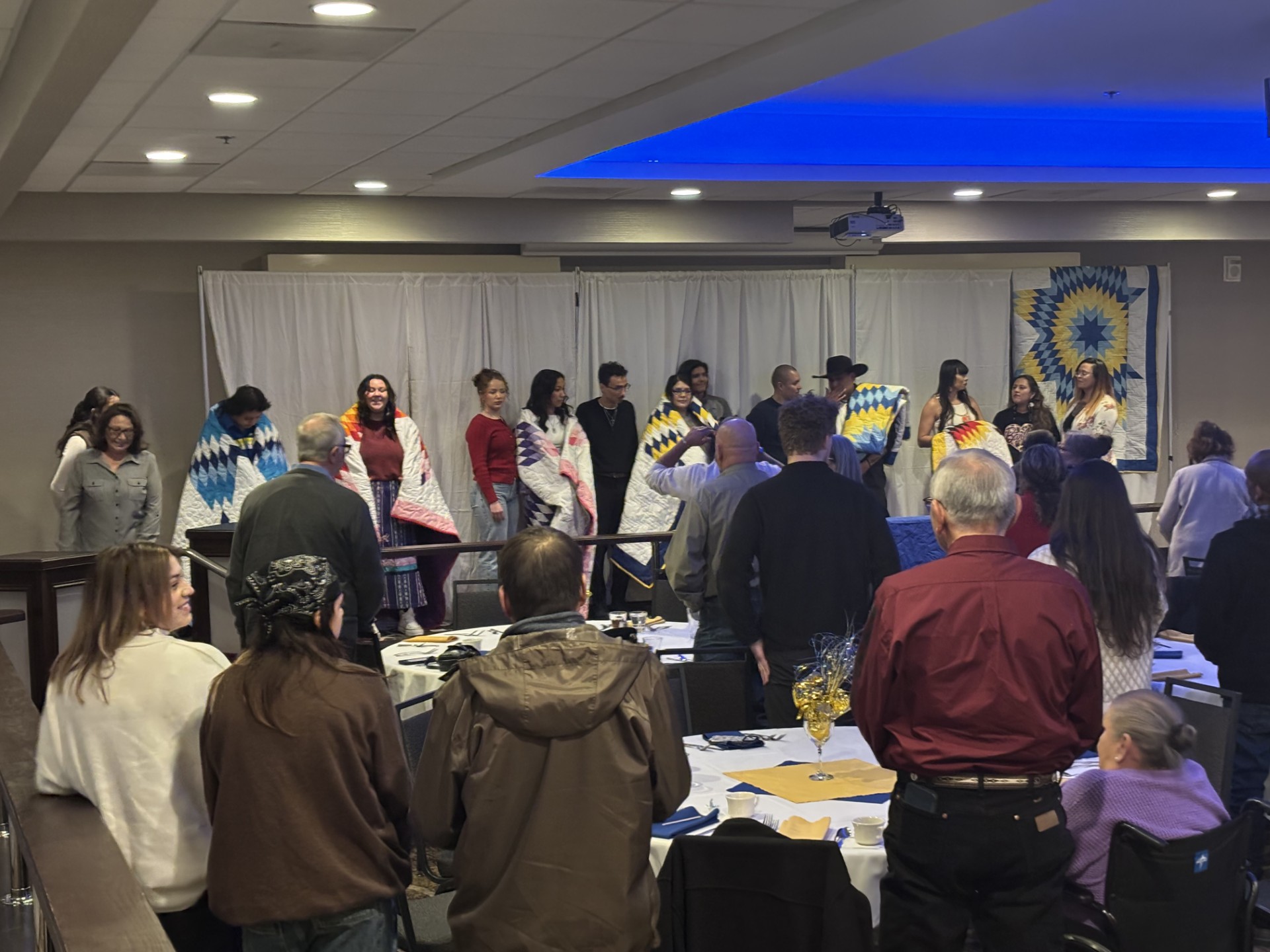 group of people in front of the room having quilts draped around their shoulders. the crowd is standing and clapping.